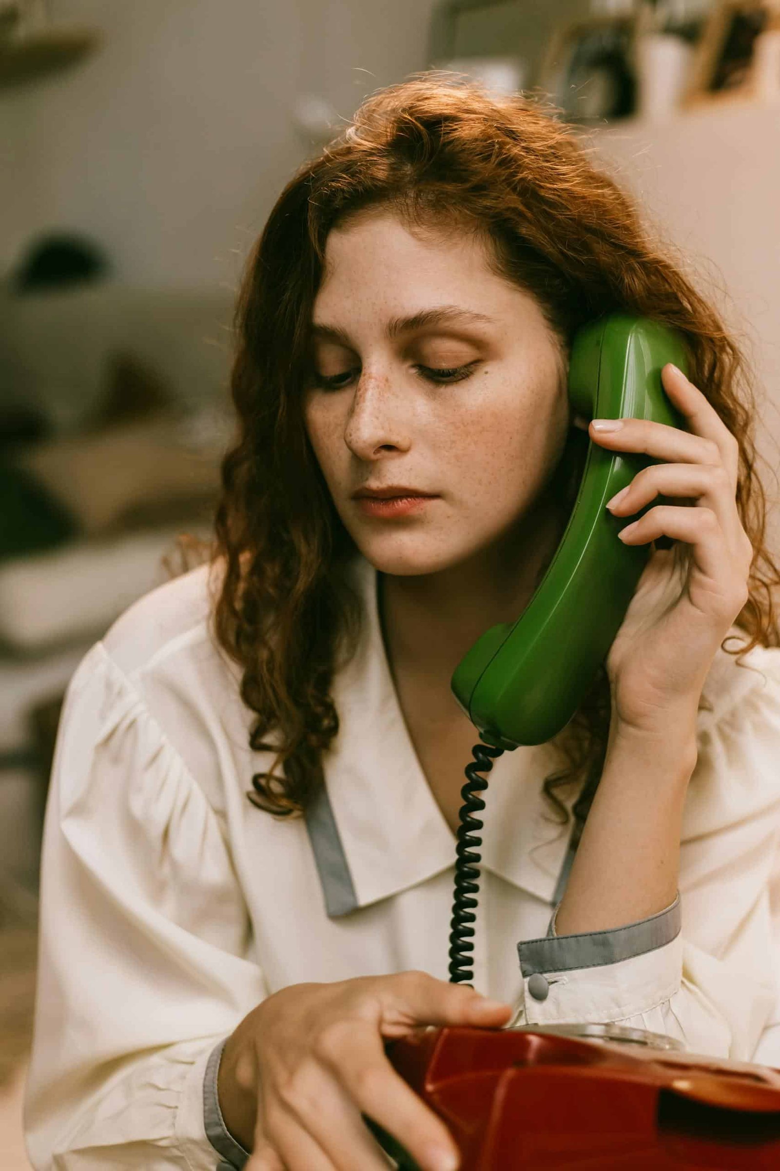 A pensive redhead woman uses a vintage green telephone, creating a nostalgic indoor scene.